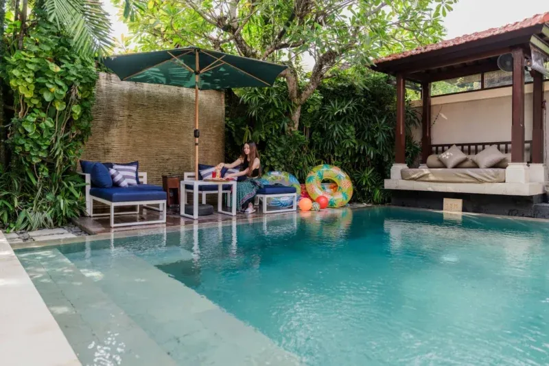 Poolside lifestyle in Seminyak, Bali — guest relaxing at navy sunloungers beside private pool, tropical garden with bamboo privacy screening, frangipani tree, bale daybed pavilion behind.