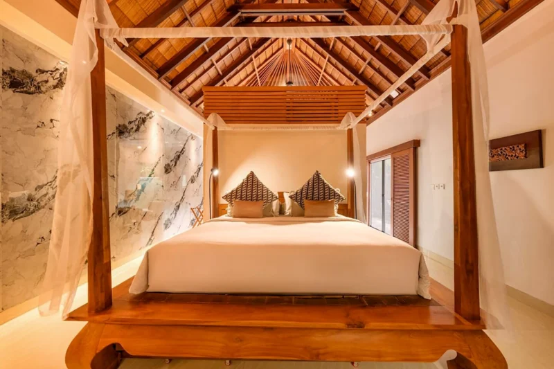 King four-poster teak canopy bed in Seminyak, Bali — straight-on view through canopy posts, floor-to-ceiling marble feature wall with spotlights, vaulted alang-alang thatched ceiling.