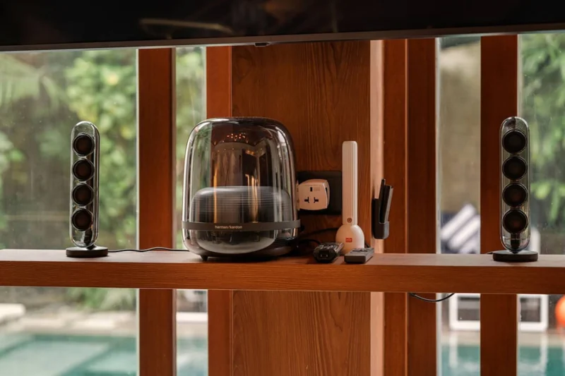 Harman Kardon premium audio system in Seminyak, Bali — SoundSticks speakers and glass subwoofer on teak TV unit shelf, private pool visible through glass doors behind.