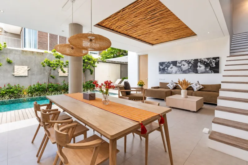 Villa Swan 01 — dining, long timber table for eight under woven rattan pendants, living room beyond