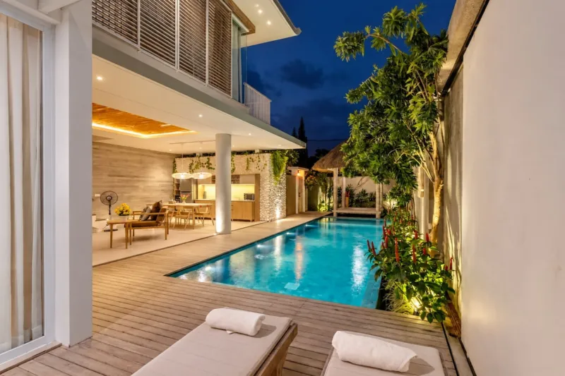 Villa Swan 01 — pool, sun loungers on the timber deck beside the lit water with the living room glowing at dusk