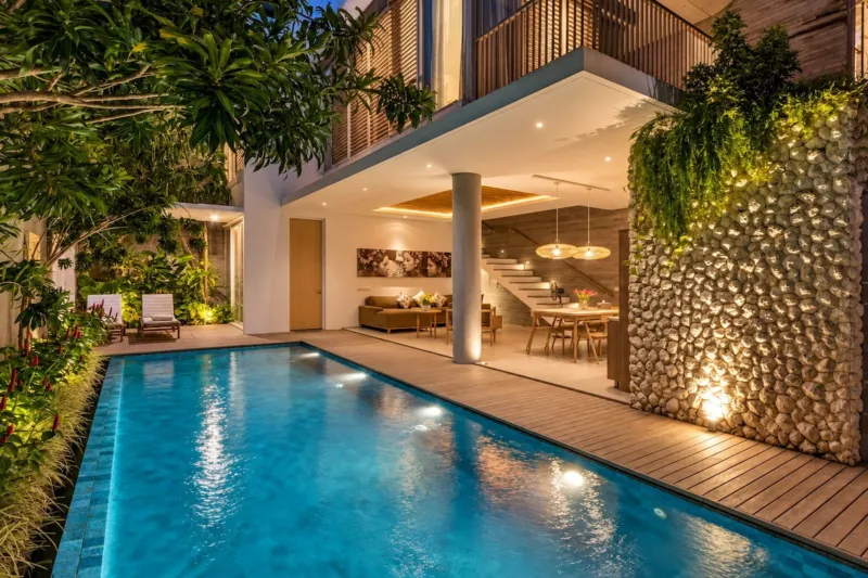 Villa Swan 01 — pool, lit at night with the open living and dining room glowing behind a stacked stone wall